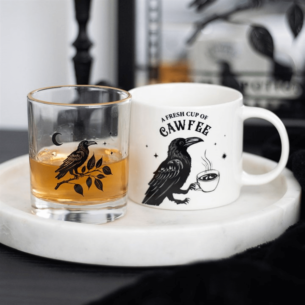 Glass with amber liquid and a white mug with a raven design and text on a marble surface.