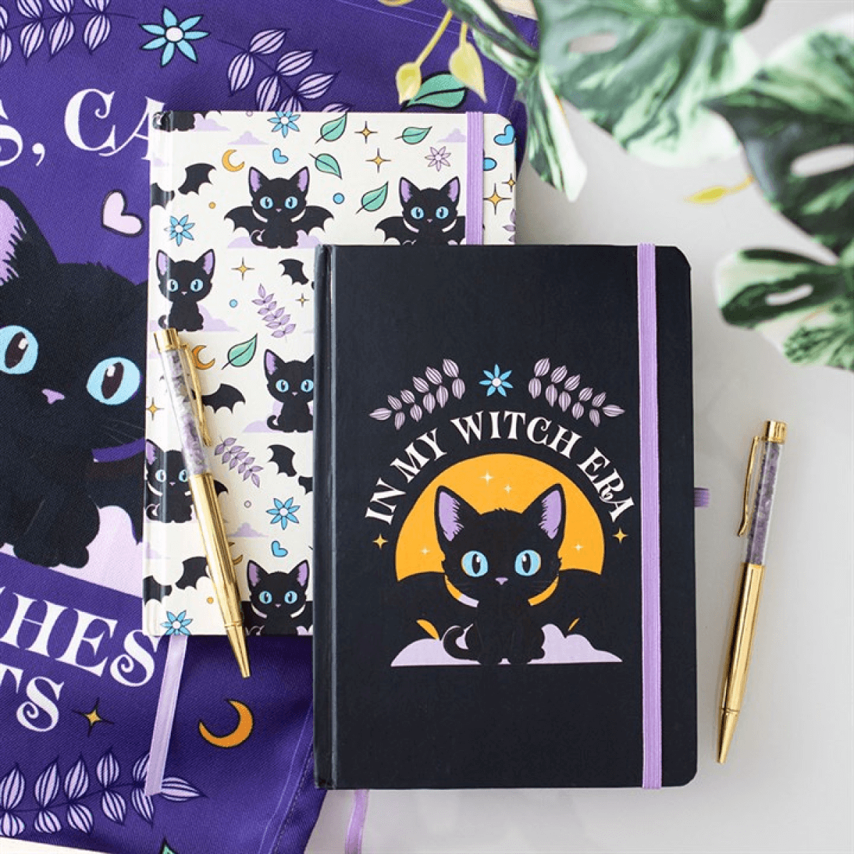 Notebooks with black cat designs on a white surface with a purple fabric and green leaves in the background.