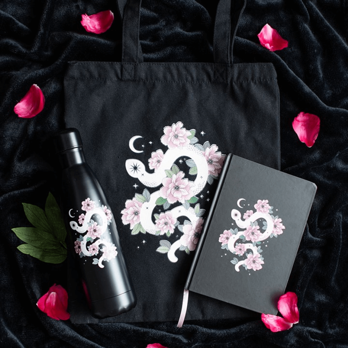 Black tote bag, water bottle, and notebook with floral dragon design on a dark fabric background with pink petals.