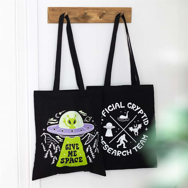 Two black tote bags with alien and UFO designs hanging on a wooden rack.