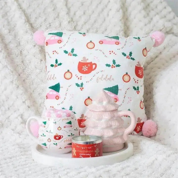 Decorative arrangement featuring a pink Christmas tree shaped mug, festive cushion, and candle on a cozy blanket.