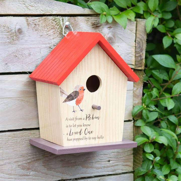 Wooden birdhouse with red roof and purple base on a wooden post with greenery.