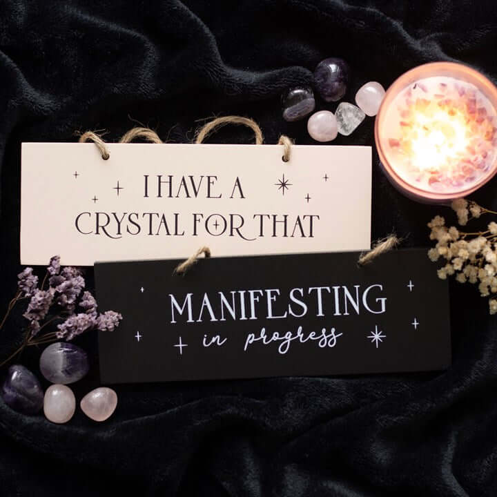 Decorative hanging sign saying 'Manifesting in progress' with a candle and crystals.
