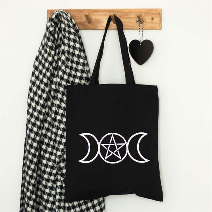 Black tote bag with a white symbol hanging on a wooden hook with a checkered scarf and heart-shaped object.