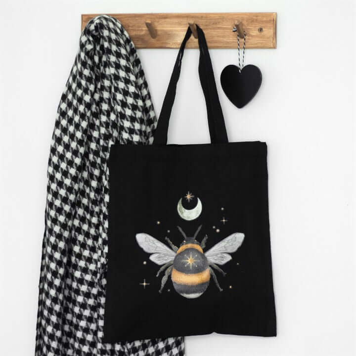 Black tote bag with bee design hanging on a wooden hook with a checkered scarf and heart-shaped object.