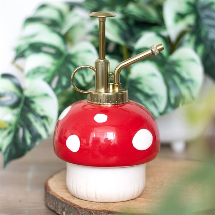 Mushroom-shaped spray bottle with red cap and white spots on a wooden surface with green plants in the background.