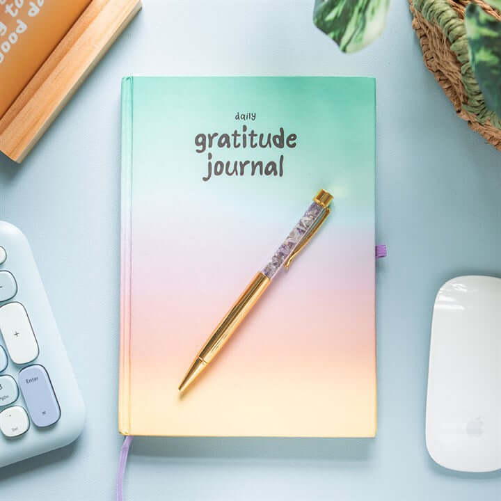 Gradient 'daily gratitude journal' on a light blue surface with a pen and computer mouse.
