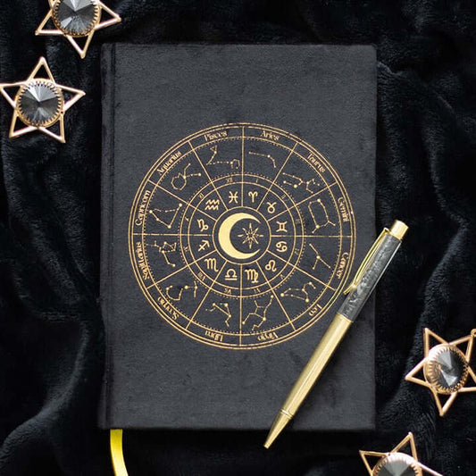 Black notebook with gold astrological design, gold pen, and decorative stars on a dark fabric background