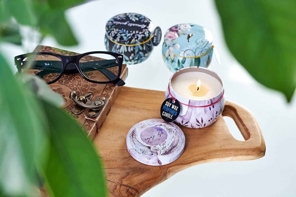 Art Tin Candle in colorful tin packaging on a wooden tray with glasses and decorative items, featuring a vanilla and musk scent.