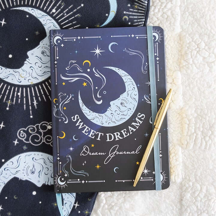 Decorative journal with celestial design and 'Sweet Dreams' text, accompanied by a pen on a textured surface.