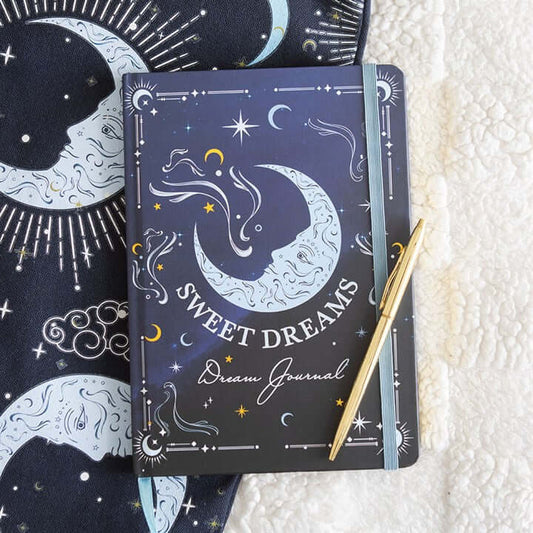 Decorative journal with celestial design and 'Sweet Dreams' text, accompanied by a pen on a textured surface.