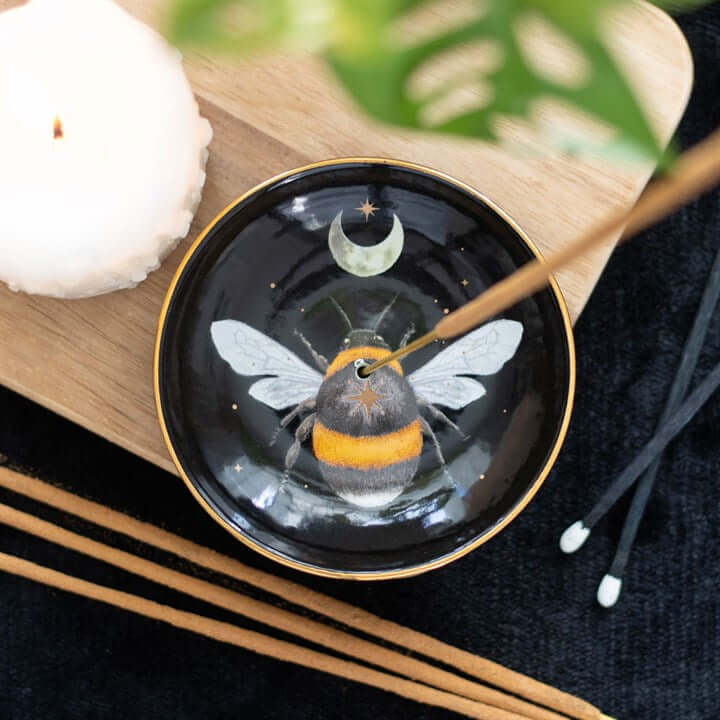 Black dish with bee design on a wooden surface with incense sticks and a candle.