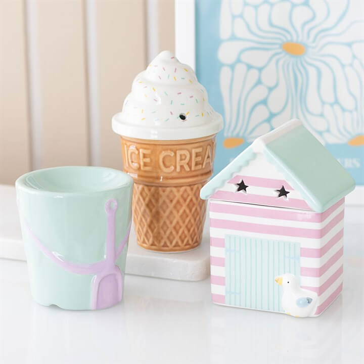 Three ceramic items including a cup, ice cream cone, and beach hut on a white surface with a floral-patterned box in the background.