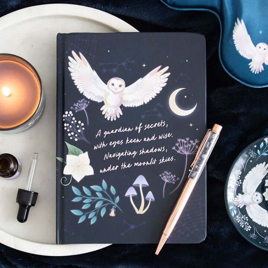 Dark notebook with owl illustration and text on a white surface with a candle and pen.