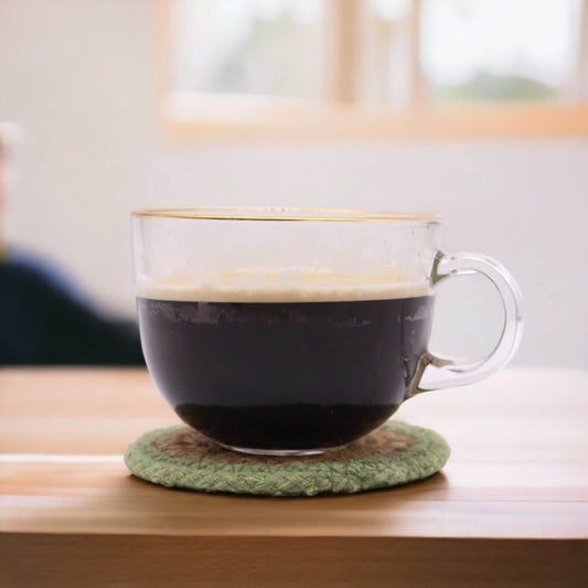 A clear coffee cup placed on a natural coaster made of jute and cotton with an olive border.