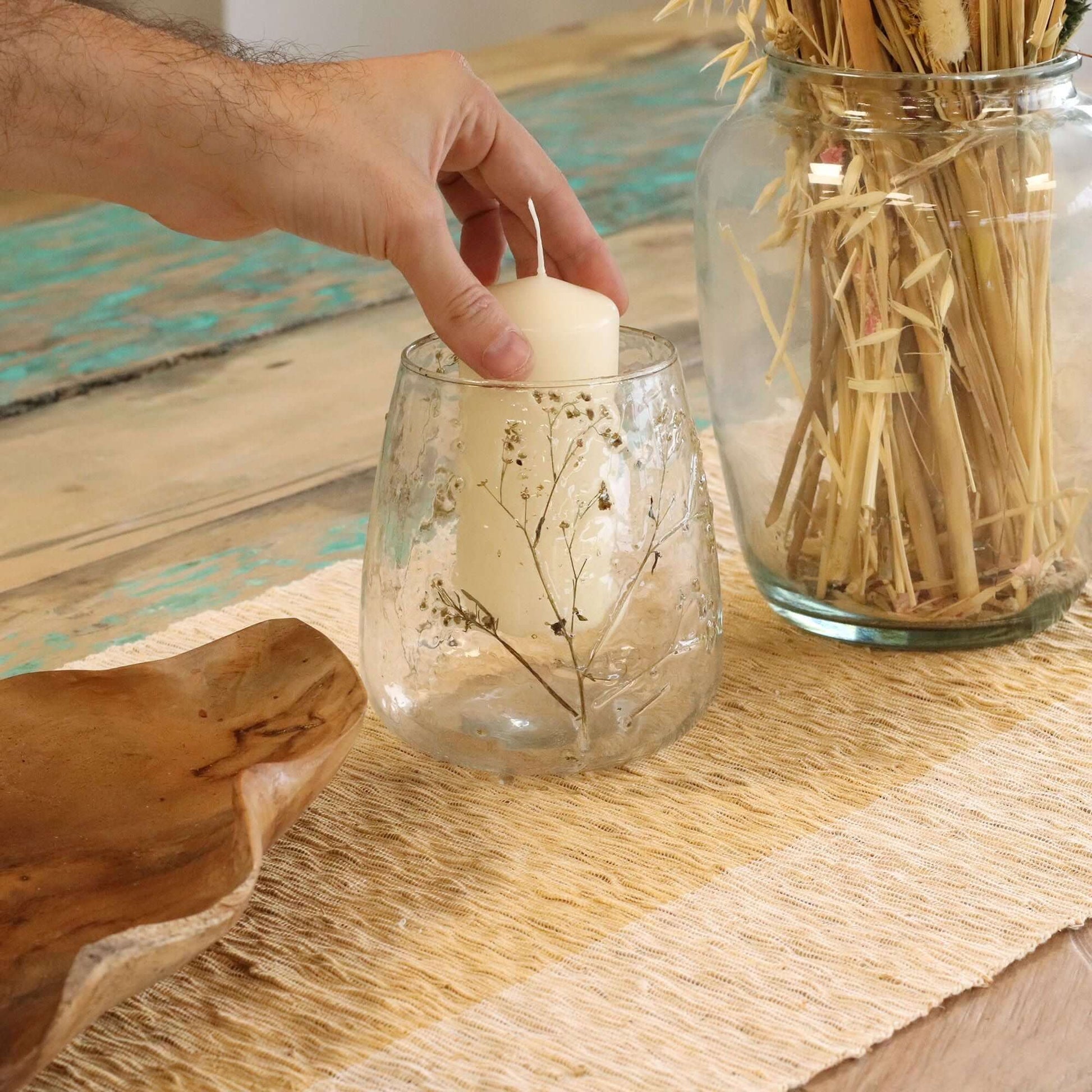 Hand placing a candle in a Natural Floral Glass vase candle holder with preserved flowers on a rustic table.