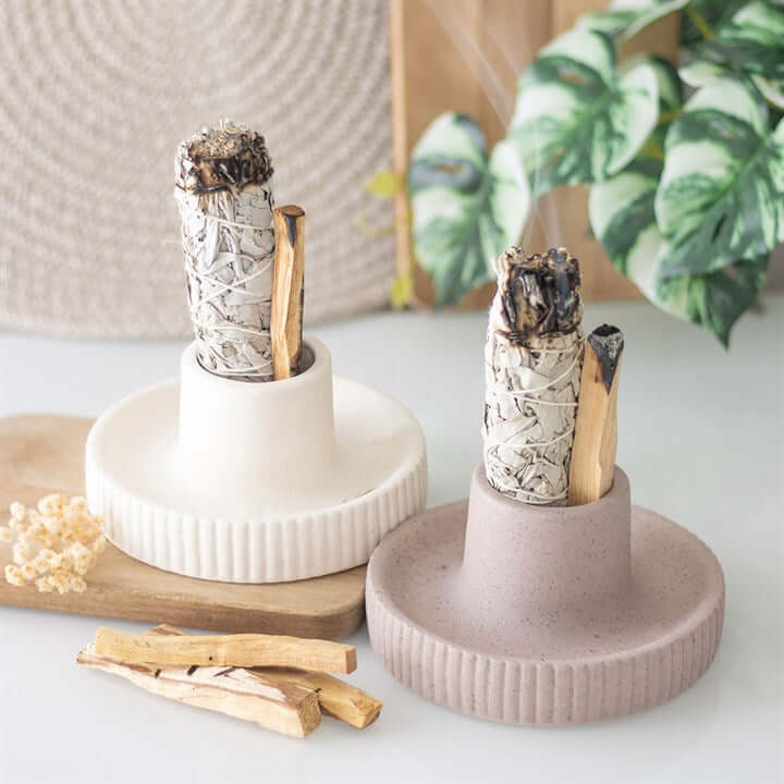 Two ceramic incense holders with incense sticks on a white surface with plants in the background