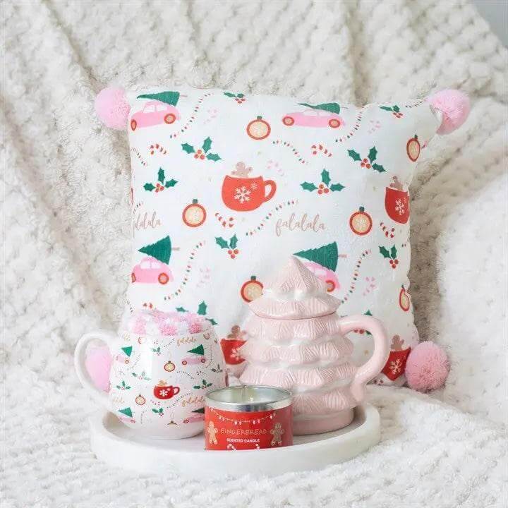 Decorative arrangement featuring a pink Christmas tree shaped mug, festive cushion, and candle on a cozy blanket.