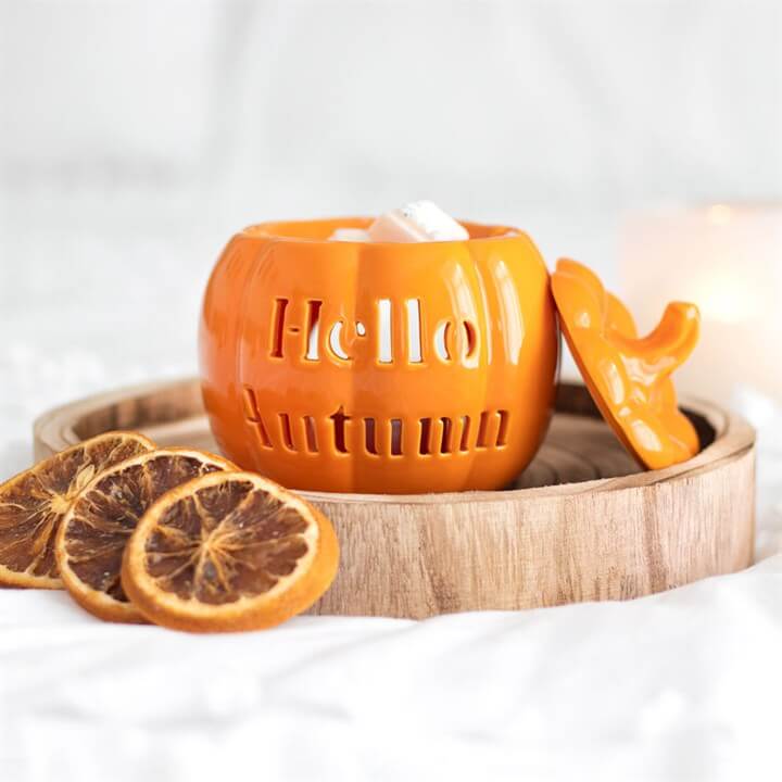 Orange ceramic pumpkin-shaped candle holder with 'Hello Autumn' text on a wooden tray with dried oranges.