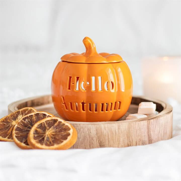 Orange pumpkin-shaped container with 'Hello Autumn' text on a wooden tray with dried oranges and candles.