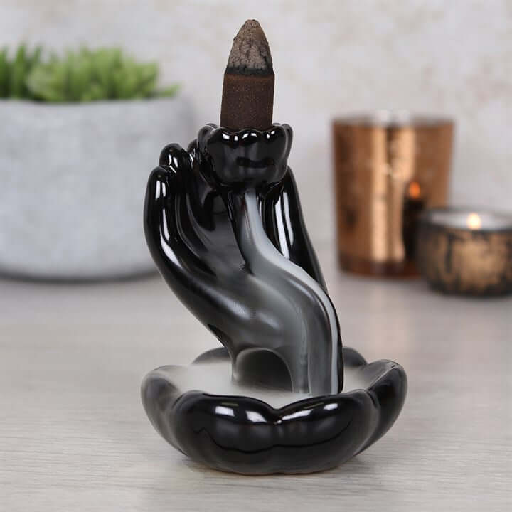 Black ceramic hand incense holder with a lit incense stick on a neutral background