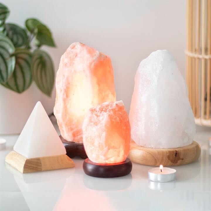 Himalayan salt lamps of various shapes and sizes on a reflective surface with a plant in the background.