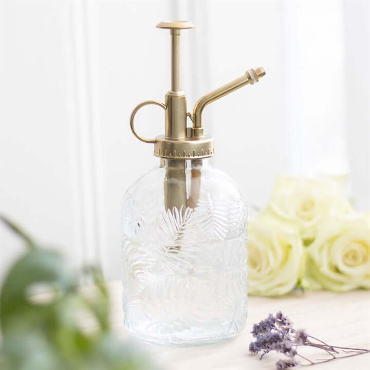 Glass spray bottle with brass nozzle on a light background with flowers and plants.
