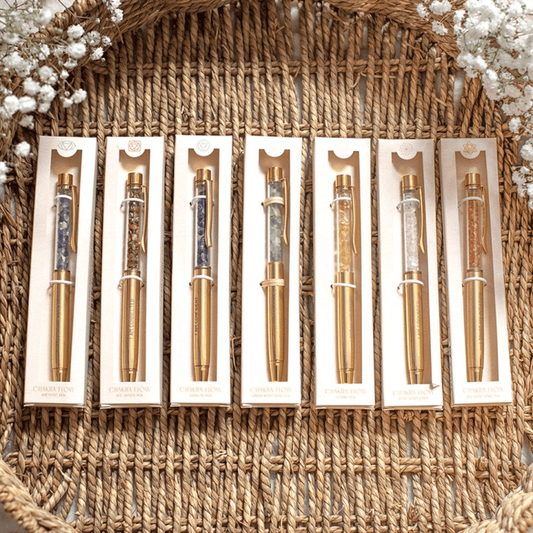 Six decorative ballpoint pens in individual white boxes, arranged side by side on a woven wicker tray with small white flowers at the corners. The pens have gold-toned barrels and clips with translucent sections showing purple, red, blue, green, and orange patterns.