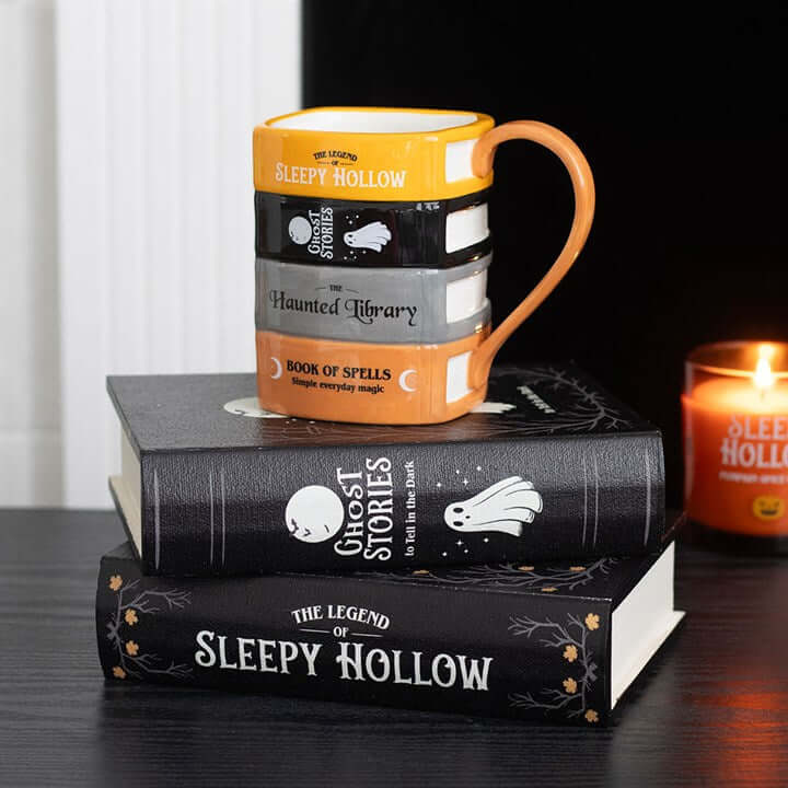 The Haunted Library Book Stack Mug on top of spooky book stack with a candle, perfect for cozy reading sessions.