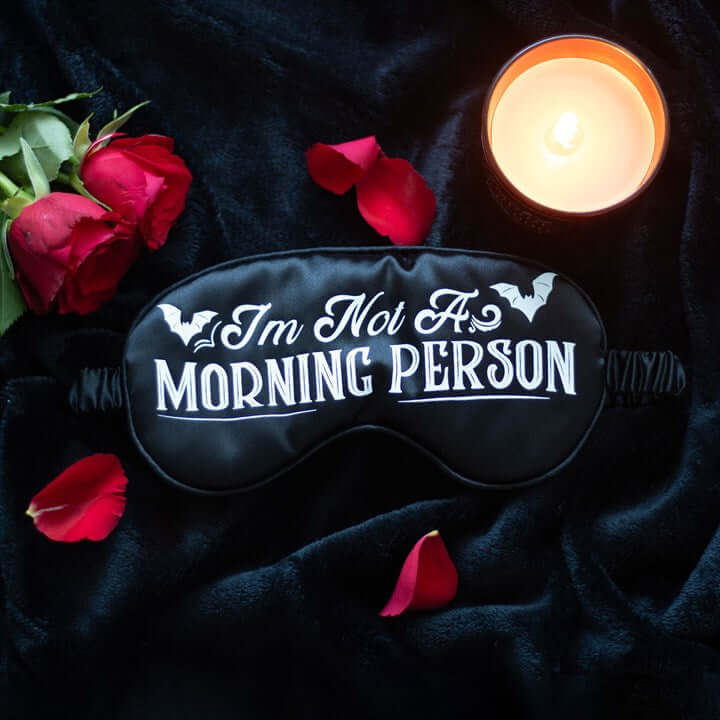Sleep mask with 'I'm Not A Morning Person' text, surrounded by red roses and a candle on dark fabric.
