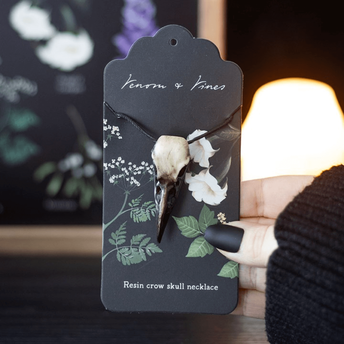 Resin crow skull necklace on a decorative card with floral designs, held by a person wearing a black knit sweater.