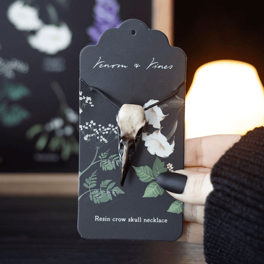 Resin crow skull necklace on a decorative card with floral designs, held by a person wearing a black knit sweater.