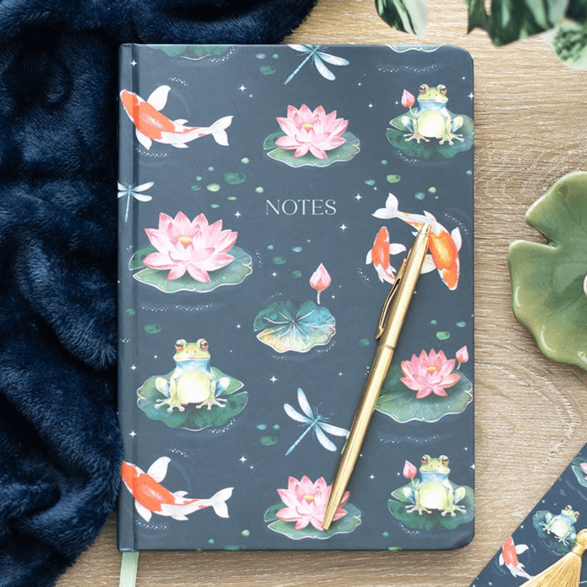 Notebook with pond-themed cover featuring frogs, fish, and lotus flowers on a wooden surface.