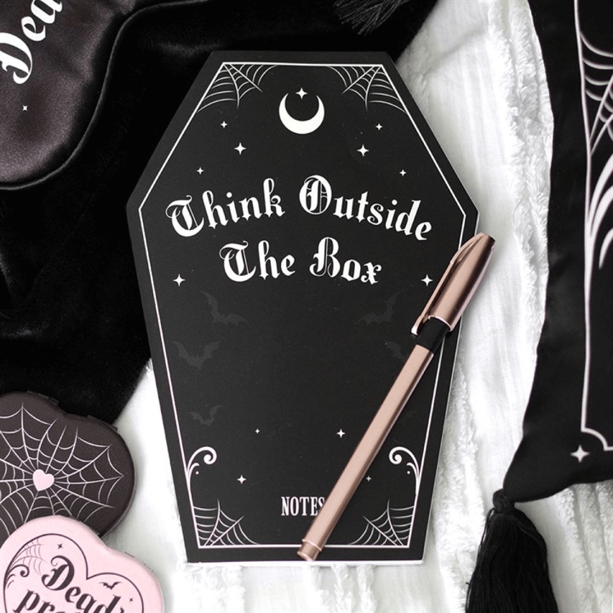 Black coffin-shaped notebook with 'Think Outside The Box' text, pen, and decorative items on a white surface.