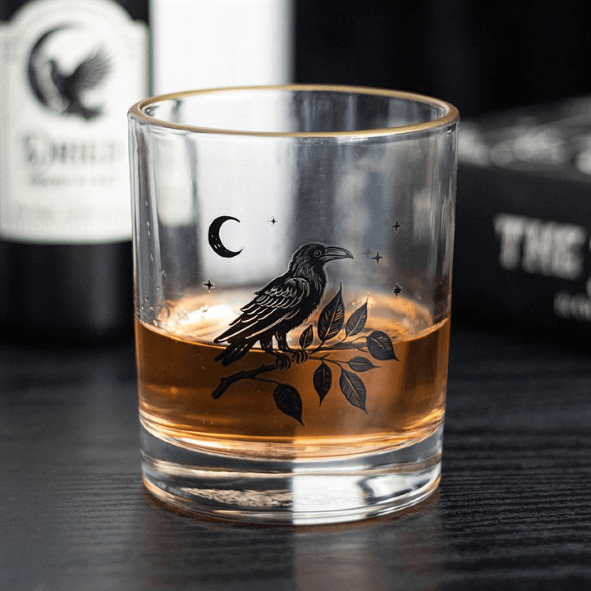 Glass with engraved raven design filled with amber liquid, blurred bottle in background
