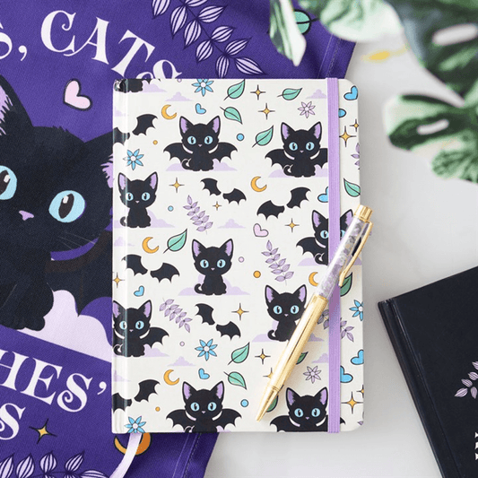 Notebook with black cat and bat pattern on a white surface with a purple book and green leaves in the background.