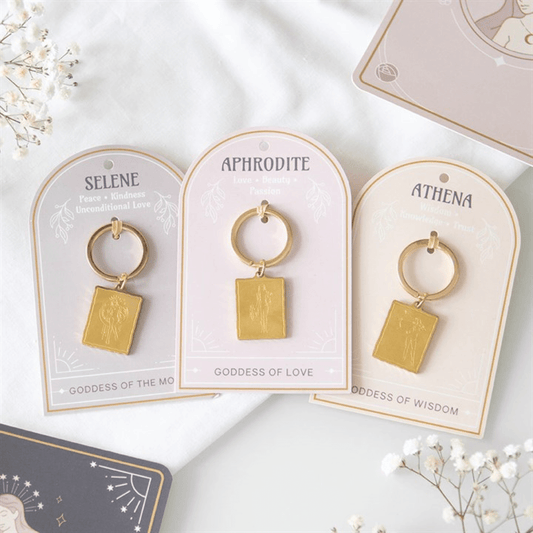 Gold keychains on name cards labeled 'Selene', 'Aphrodite', and 'Athena' on a white background with flowers.