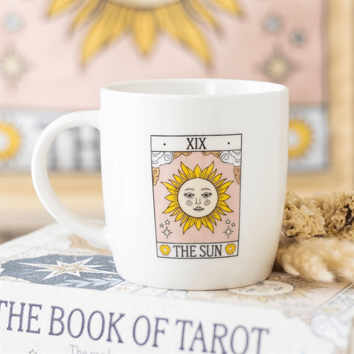 White mug with 'The Sun' tarot card design on a surface with books and decorative items.