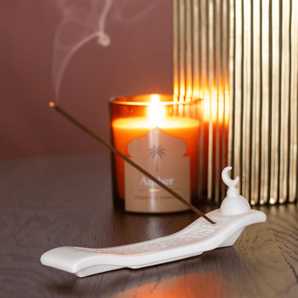 Incense stick burning on a white ceramic holder with a lit candle in the background.