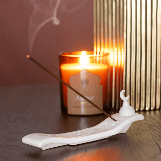 Incense stick burning on a white ceramic holder with a lit candle in the background.