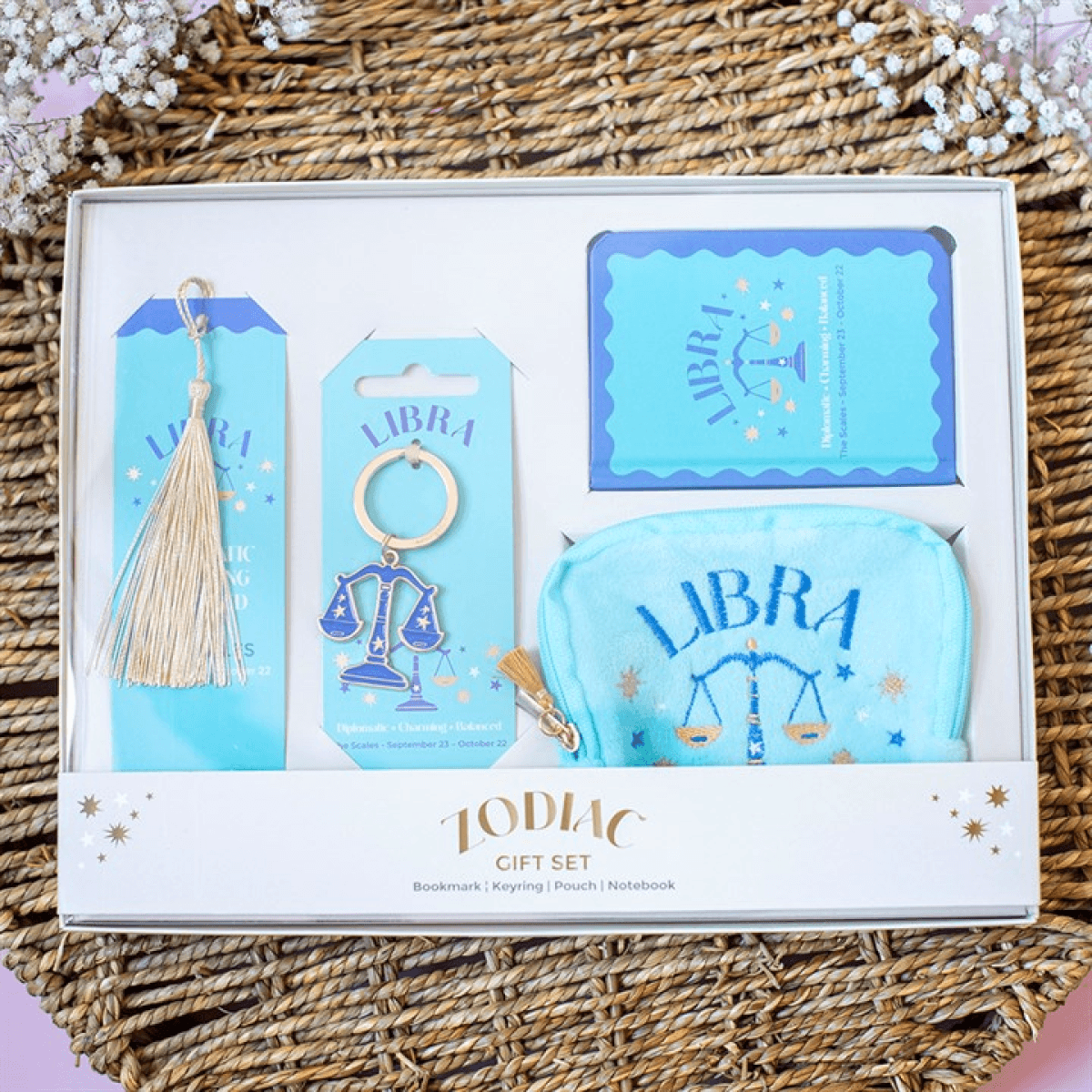 A Libra-themed gift set in a white box, featuring a light blue bookmark with a gold tassel, a purple Libra keychain, a blue card, and a matching light blue pouch, all displayed on a woven wicker mat.