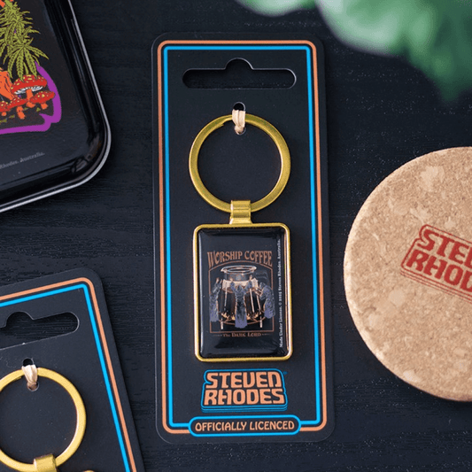 Gold keychain with 'Steven Rhodes' branding on a black card, surrounded by other branded items.
