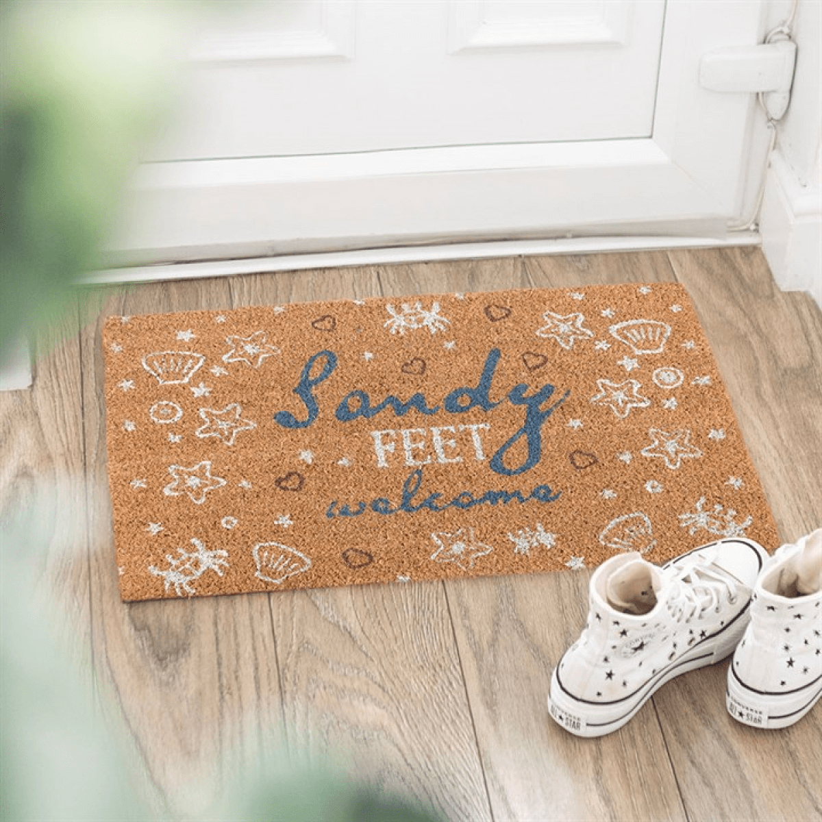 Doormat with 'Sandy Feet Welcome' text on a wooden floor with white sneakers.